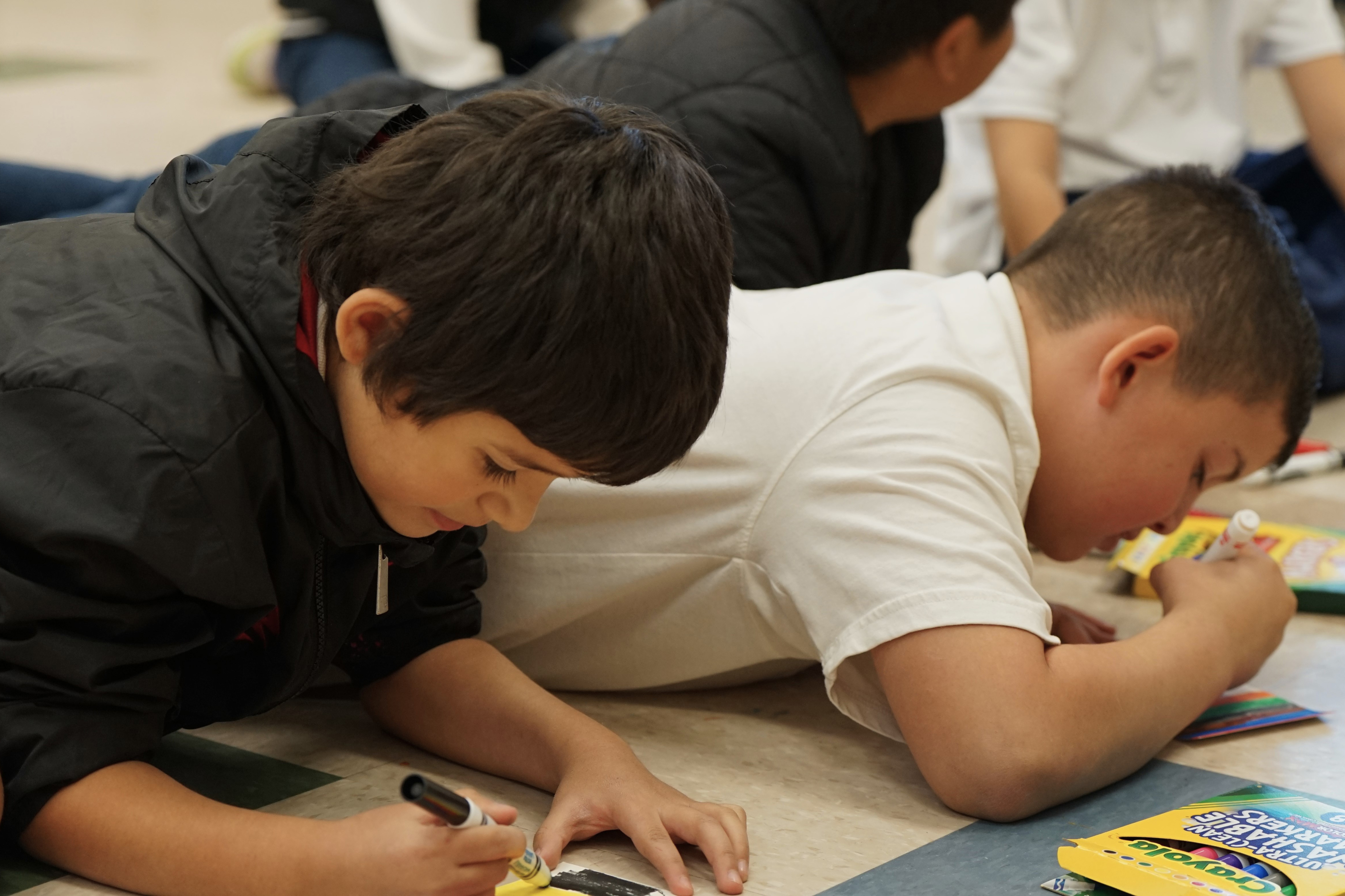 Two boys lay on the floor drawing their rug designs