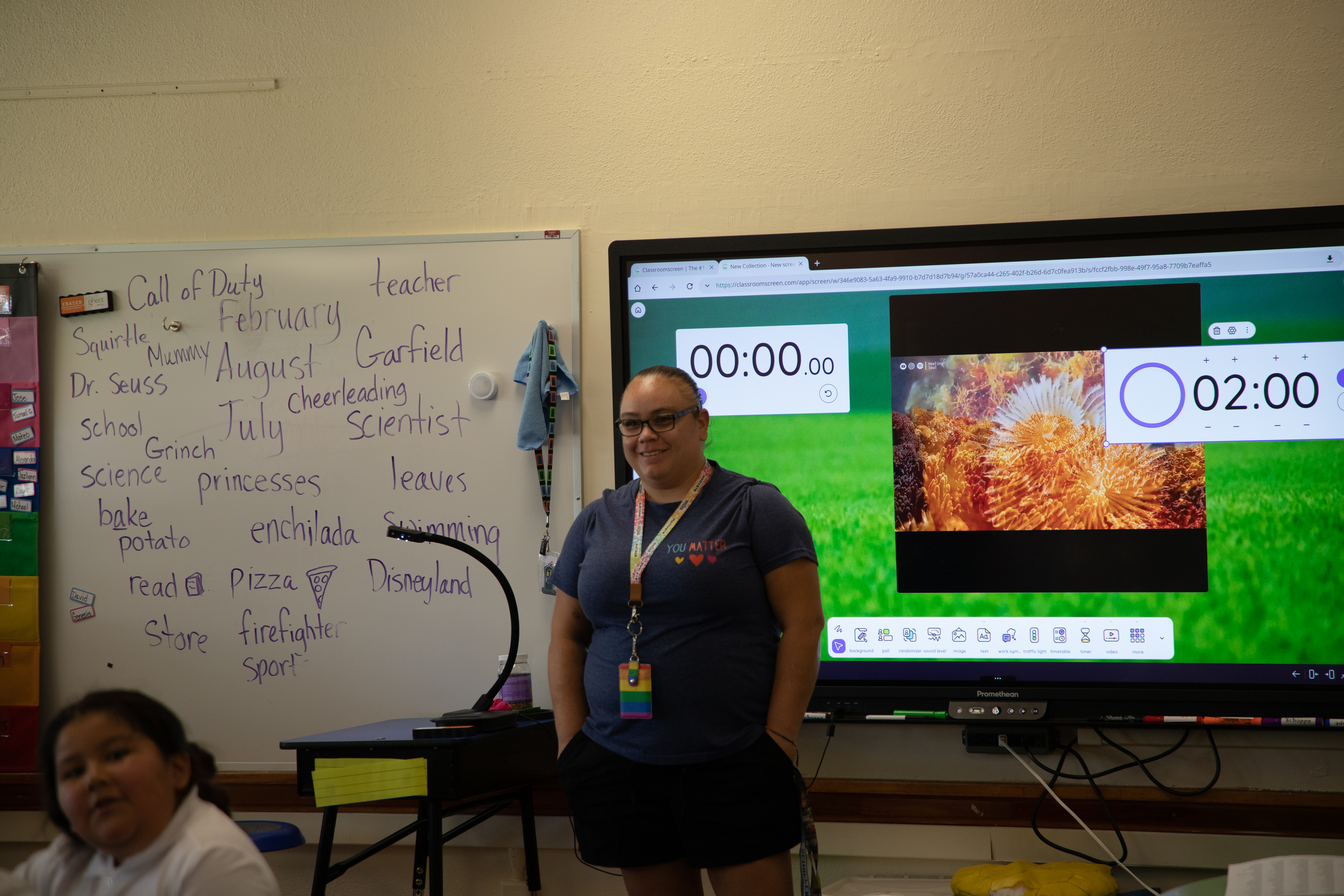 A teacher stands at the front of her classroom smiling at her students