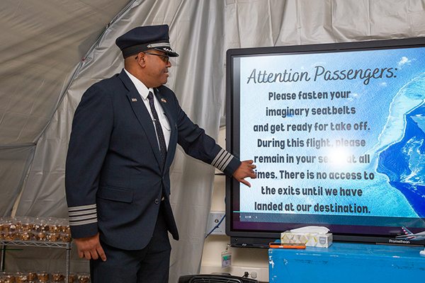 A man dressed as a pilot points out the safety message to students on the screen